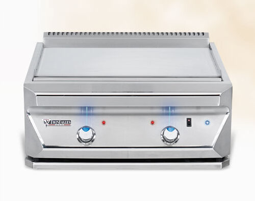 A stainless steel commercial salamander broiler with two blue-flamed heating elements and control knobs, set against a soft neutral background near a fireplace.