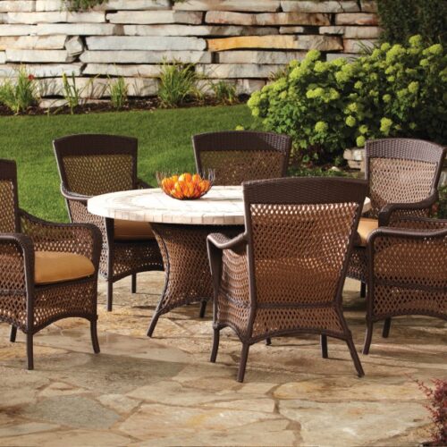 Outdoor patio featuring a round stone table and six wicker chairs, set on a patterned tile surface with a lush garden and stone fire pit in the background.