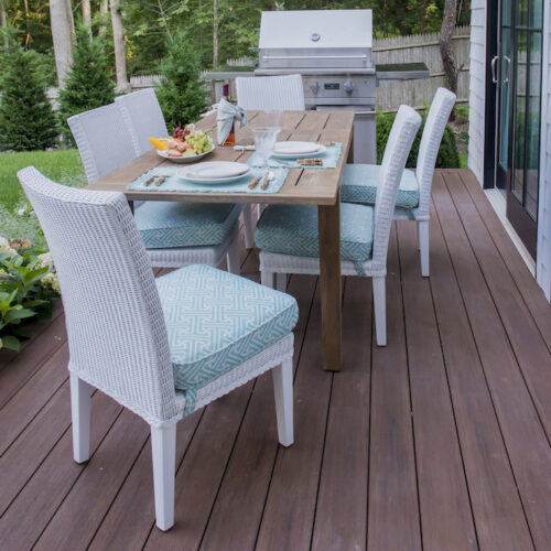 An outdoor dining area on a wooden deck with a table set for four, white wicker chairs with blue cushions, surrounded by lush greenery and a fire pit nearby.