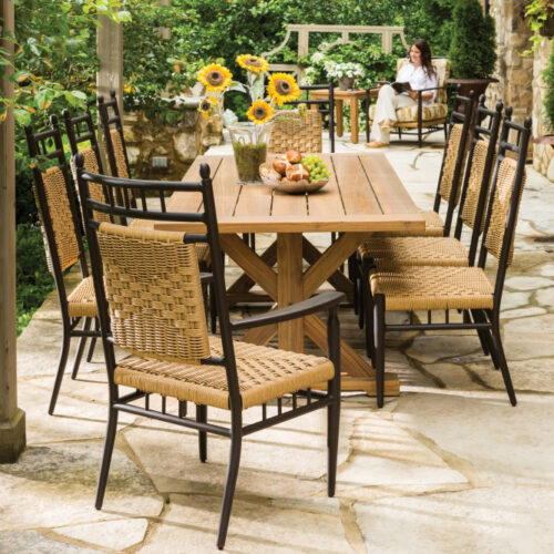 An outdoor dining area with a wooden table and wicker chairs, set with fruit and flowers. In the background, a woman sits relaxed in a chair, surrounded by lush greenery and a cozy fire