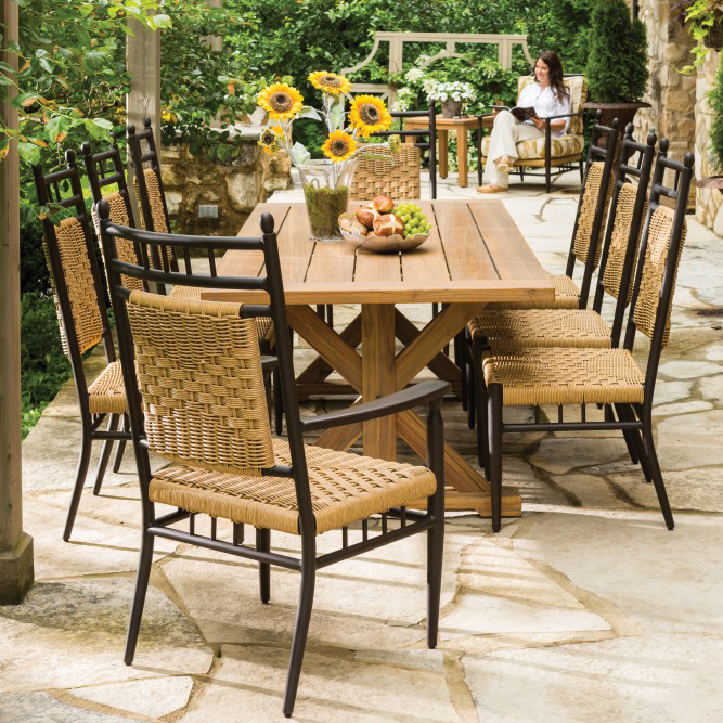 An outdoor dining area with a wooden table and wicker chairs, set with fruit and flowers. In the background, a woman sits relaxed in a chair, surrounded by lush greenery and a cozy fire