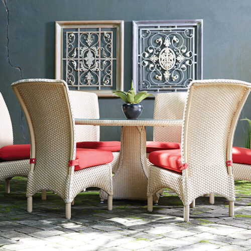 Outdoor patio setting with a rectangular table and six high-back wicker chairs with red cushions, positioned on a stone floor against a wall with a fireplace and two decorative metal panels.