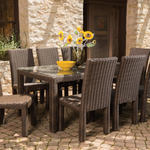 An outdoor dining area featuring a rattan table and chairs set on a cobblestone patio, with sunflowers in a yellow bowl centerpiece and a rustic stone building with a fire pit in the
