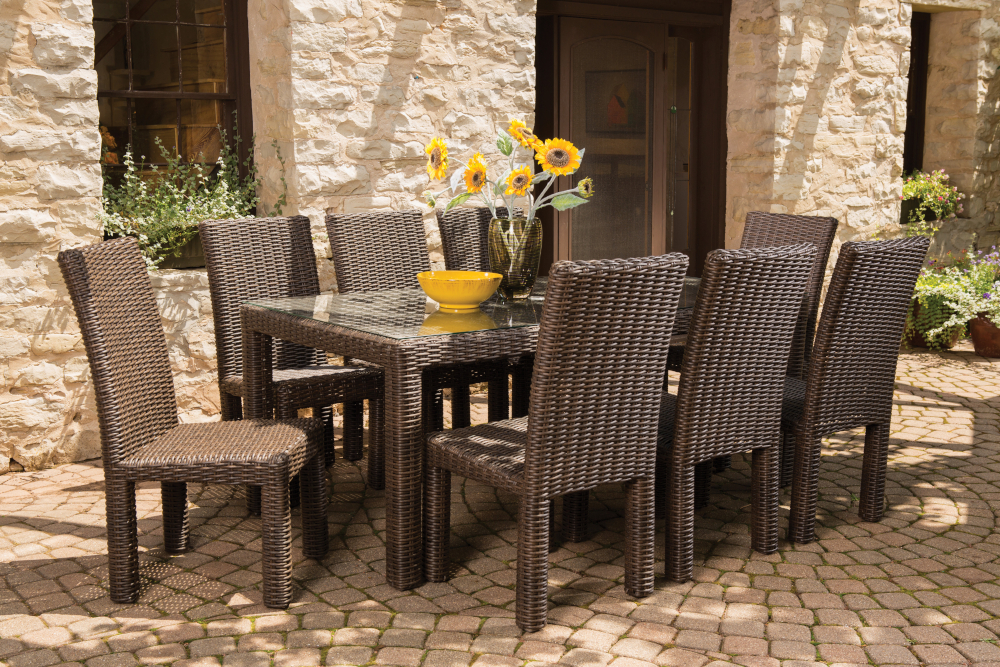 An outdoor dining area featuring a rattan table and chairs set on a cobblestone patio, with sunflowers in a yellow bowl centerpiece and a rustic stone building with a fire pit in the
