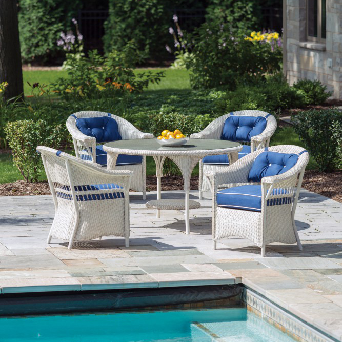 A patio set with four white wicker chairs featuring navy blue cushions around a round table, beside a pool, in a garden setting with a fire pit nearby. A bowl of lemons is on the