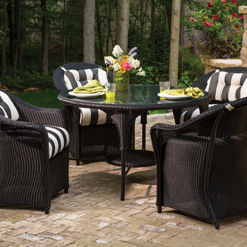 Outdoor patio set with a black wicker table and four matching chairs, striped cushions, and a set table with plates and flowers, in a garden with a stone wall, greenery, and an insert