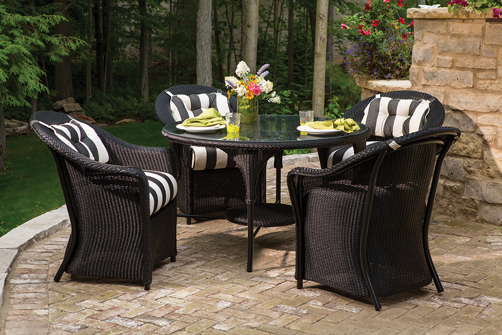 Outdoor patio set with a black wicker table and four matching chairs, striped cushions, and a set table with plates and flowers, in a garden with a stone wall, greenery, and an insert