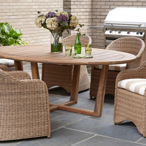 A cozy outdoor patio featuring a round wooden table and four wicker chairs with cushions, near a grill and potted plants, set against a brick wall with an inserted fireplace.