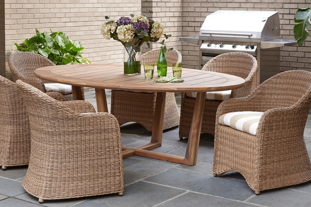 A cozy outdoor patio featuring a round wooden table and four wicker chairs with cushions, near a grill and potted plants, set against a brick wall with an inserted fireplace.