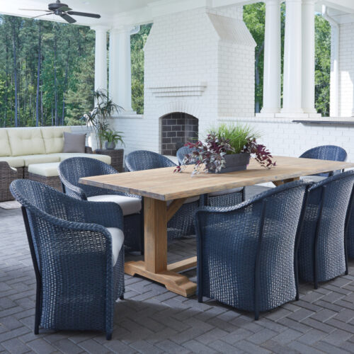 A luxurious outdoor patio with a wooden dining table and blue wicker chairs, white brick columns, a fire pit, and lush forest views in the background.