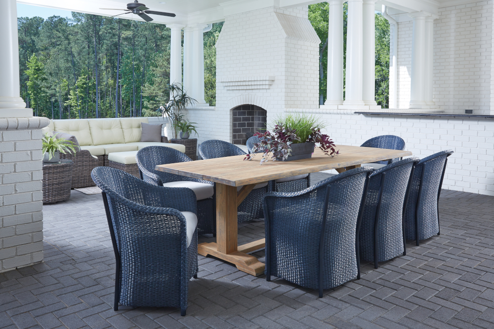 A luxurious outdoor patio with a wooden dining table and blue wicker chairs, white brick columns, a fire pit, and lush forest views in the background.