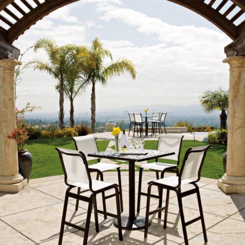 An elegant outdoor dining area under a curved wooden pergola, with a table set for four and a nearby fire pit, overlooking a scenic, lush landscape under a clear sky.