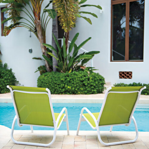 Two green lounge chairs on a tiled patio beside a serene pool, with lush palm trees and a modern fireplace visible in the background.