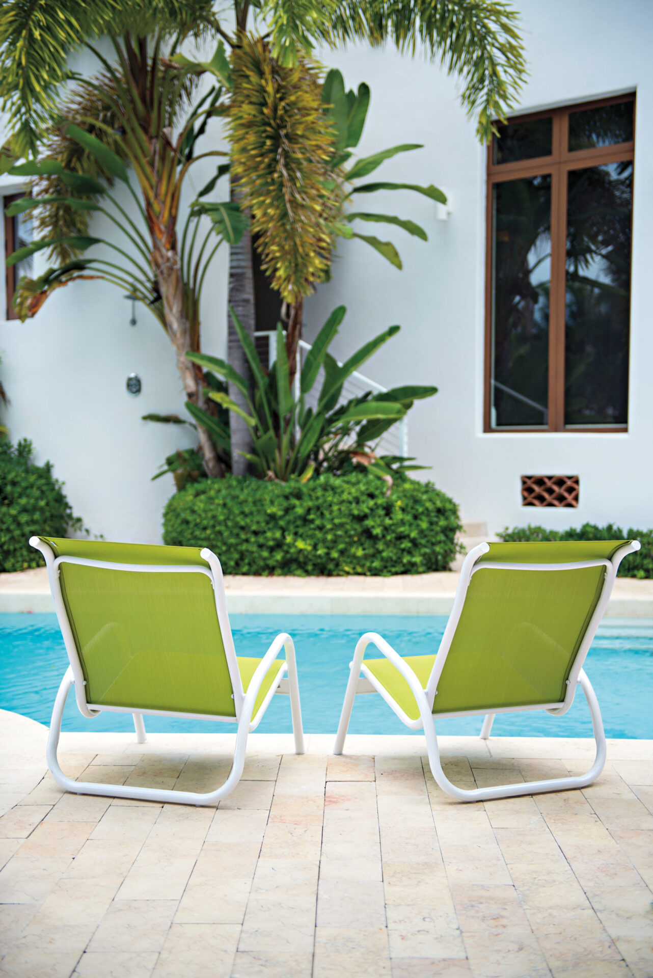 Two green lounge chairs on a tiled patio beside a serene pool, with lush palm trees and a modern fireplace visible in the background.