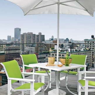 A rooftop patio with a white table and bright green chairs under a large white umbrella, overlooking a cityscape with multiple buildings under a clear sky, enhanced by a modern fireplace insert.