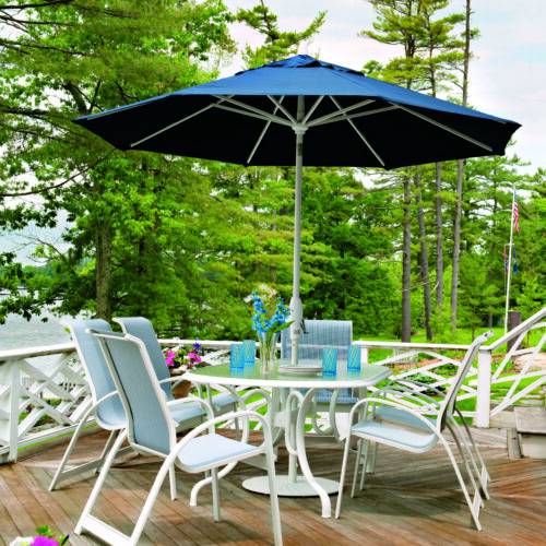 A scenic outdoor setting with Primera Sling white patio furniture and a large blue umbrella on a wooden deck, overlooking a lush forest and lake. A serene environment for relaxation or dining.