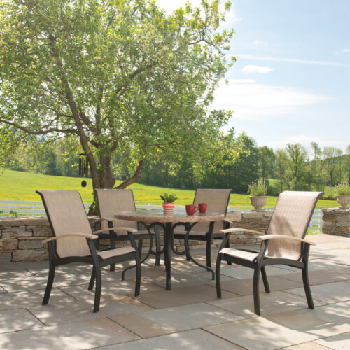 A Belle Isle Sling Collection patio set with four chairs around a table under a lush tree, overlooking a grassy field on a sunny day.
