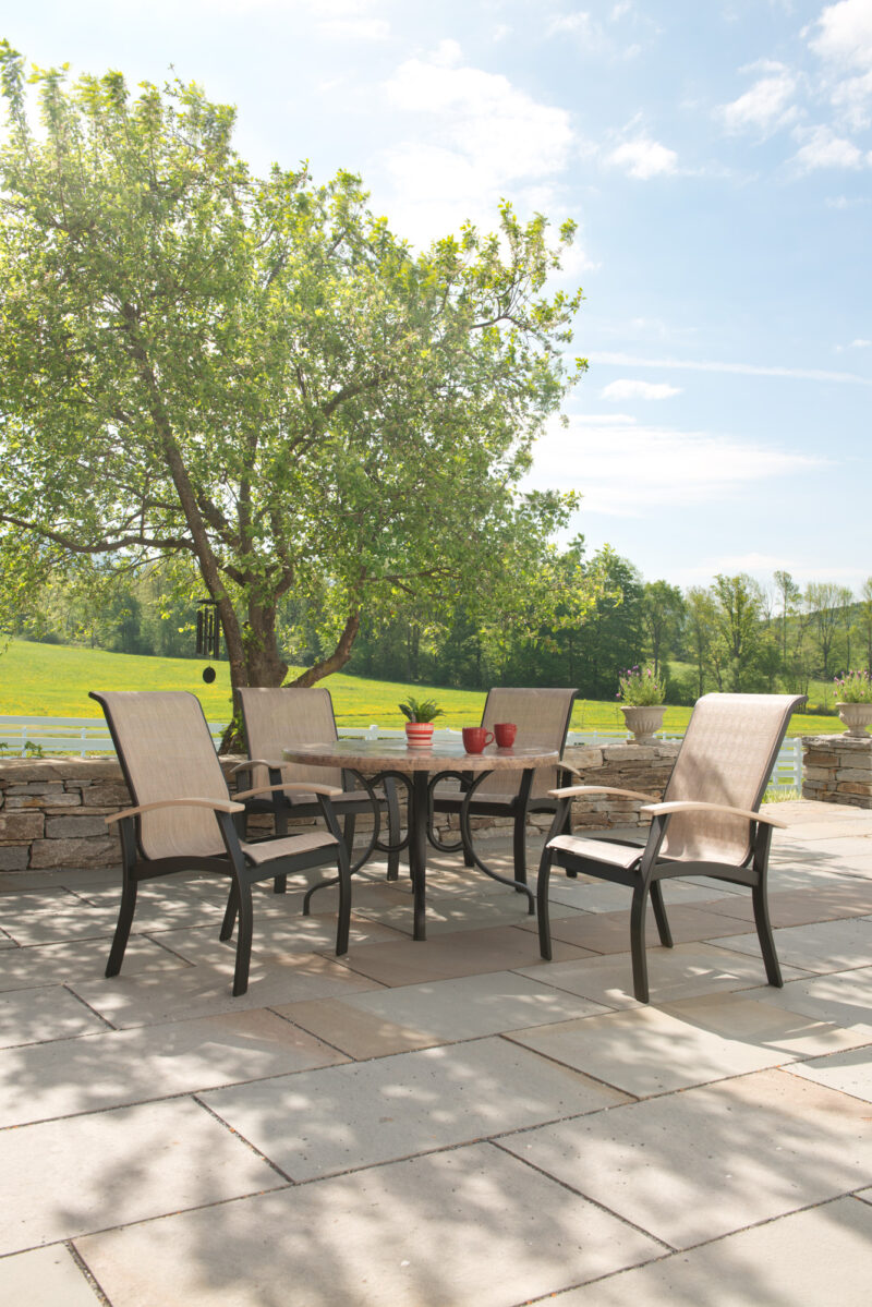 A Belle Isle Sling Collection patio set with four chairs around a table under a lush tree, overlooking a grassy field on a sunny day.