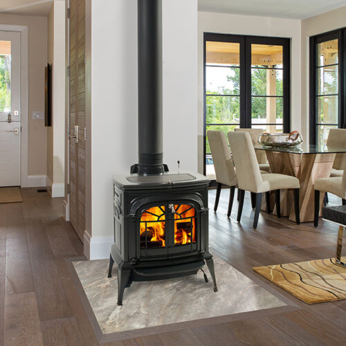 Modern living room with a lit fireplace stove, hardwood floors, large windows, a dining table set, and a view of greenery outside.