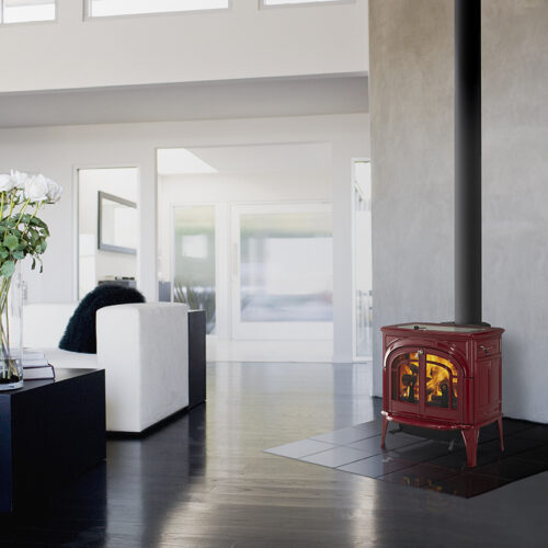 A sleek, modern living room featuring a vibrant red wood stove on a black tiled platform, with white walls and large windows shedding natural light.