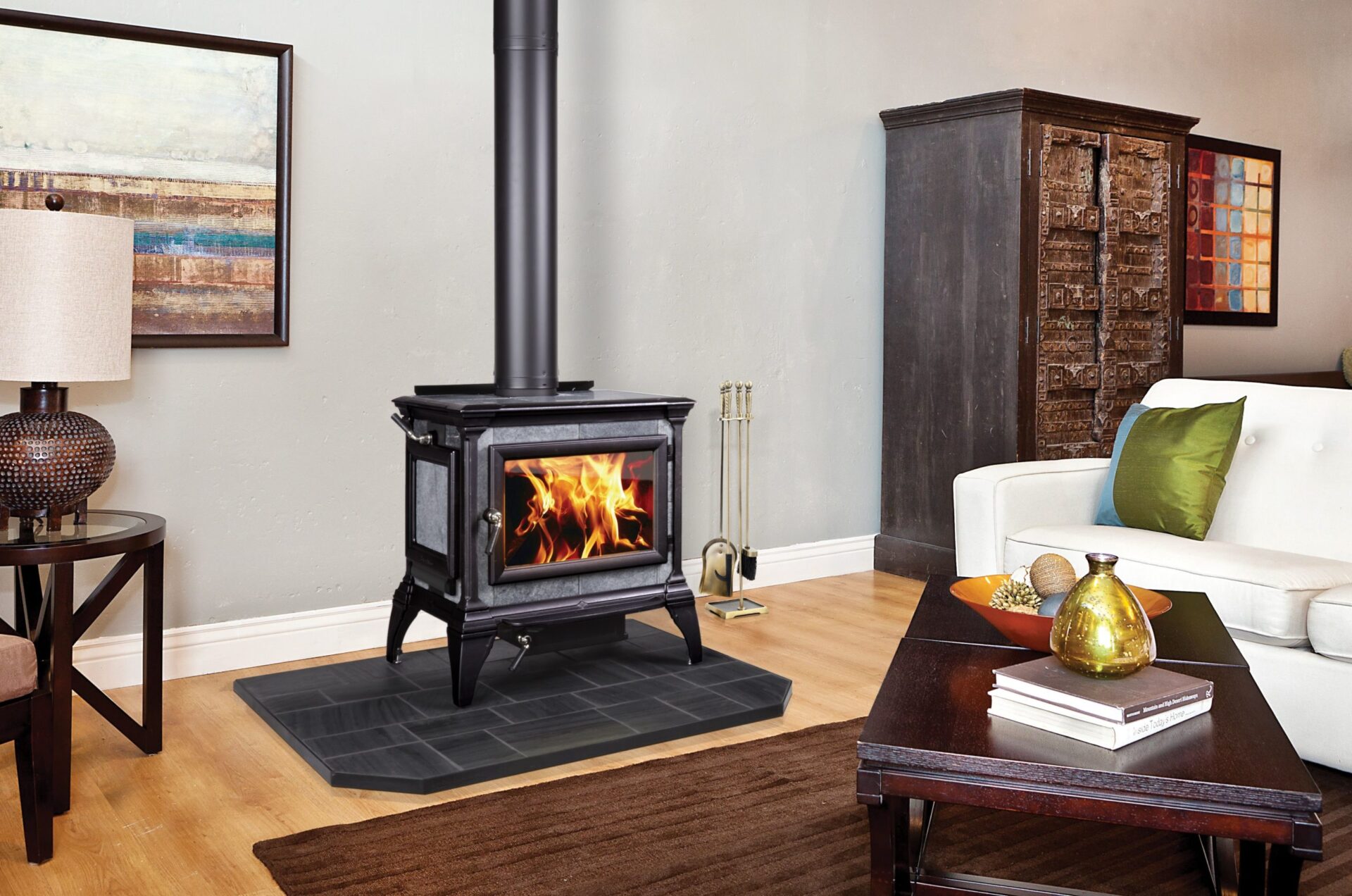A cozy living room featuring a wood-burning stove with visible flames, a white sofa with green pillows, an elegant coffee table, and colorful wall art.