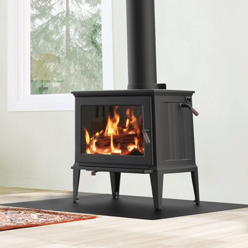A modern wood stove with a visible fire through its glass door, installed in a room featuring a white wall, window, and a Weber Grills patterned rug.