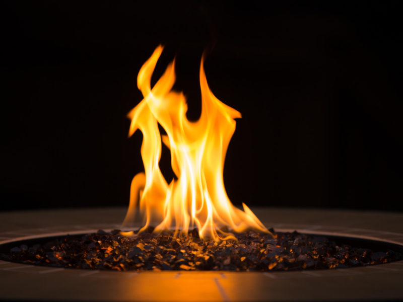 a close-up of a round, gas fire pit with red and orange flames in an outdoor living space at night