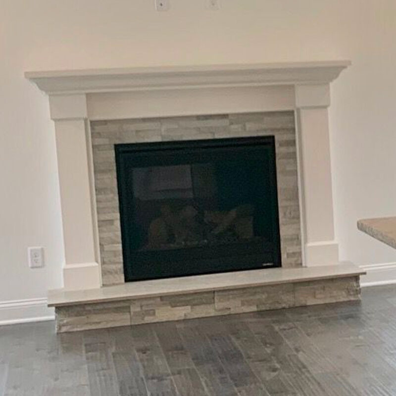 A modern fireplace with a beige mantel and surround, featuring stacked stone details, set against a plain white wall in a room with light hardwood flooring.