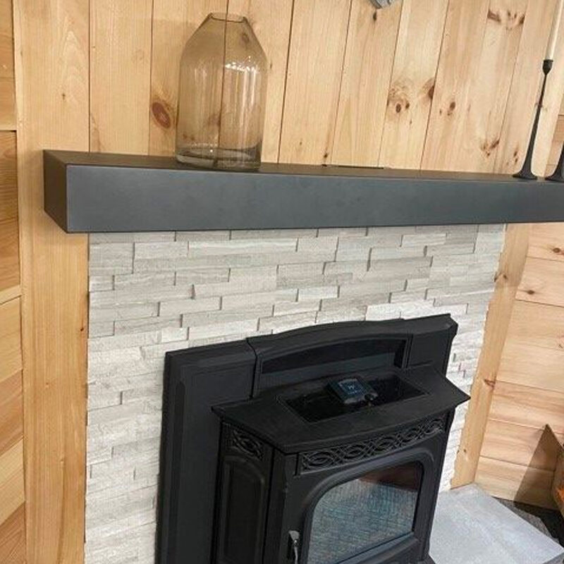 A modern, black wood-burning stove with glass door, situated against a white brick-style wall, under a dark shelf with a large clear glass jar on it. a wooden background enhances the rustic ambiance.