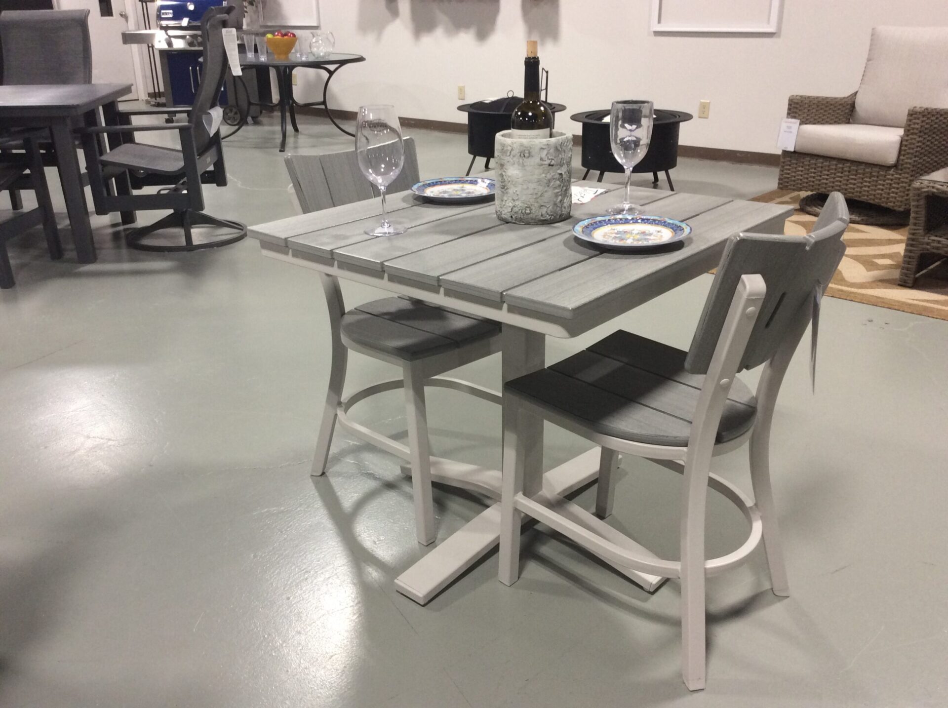 A modern dining table set with grey and white finishes, featuring two chairs and a bench, elegantly set with plates and wine glasses, displayed in a furniture showroom.
