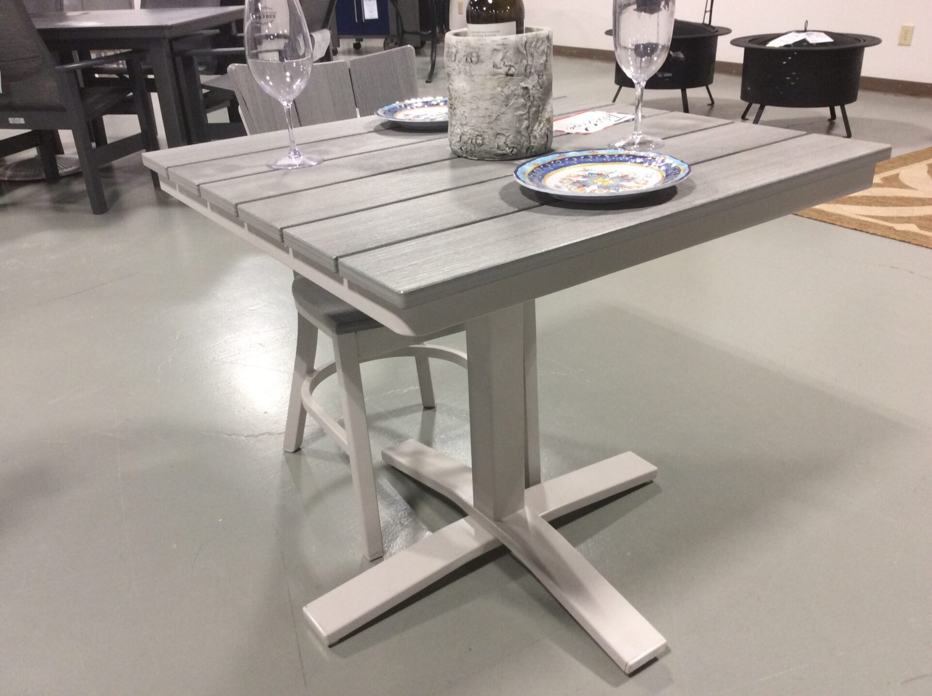 A modern dining table set in a furniture showroom, featuring a gray wood finish with a unique x-shaped base. the table is set with blue patterned plates and clear wine glasses.