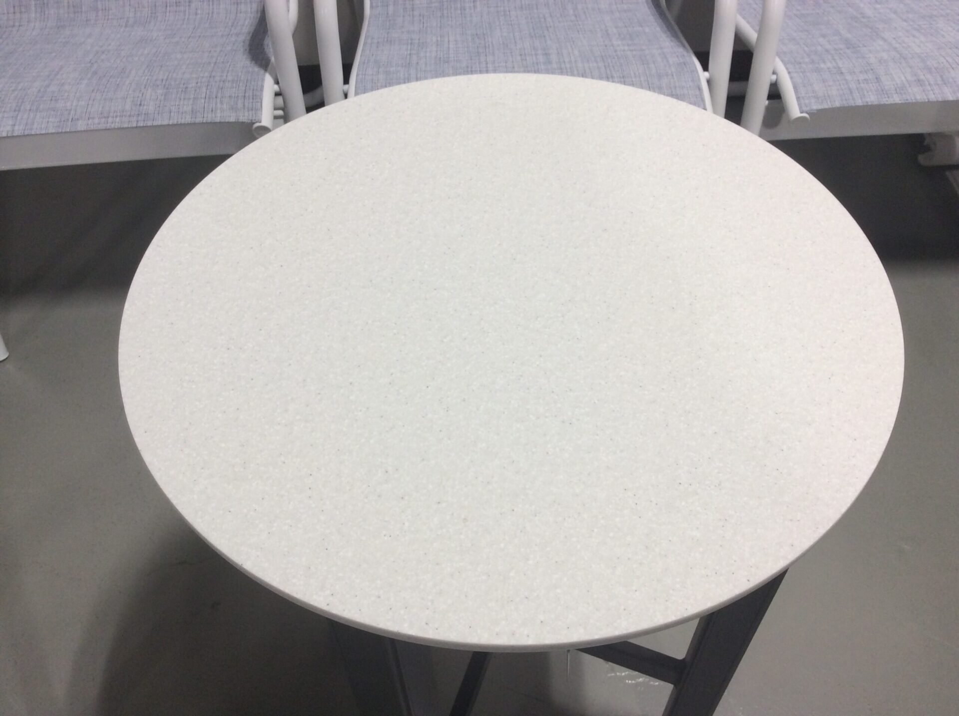 A close-up view of a round, white speckled table with metal legs, surrounded by white chairs with metal frames, set on a gray floor.