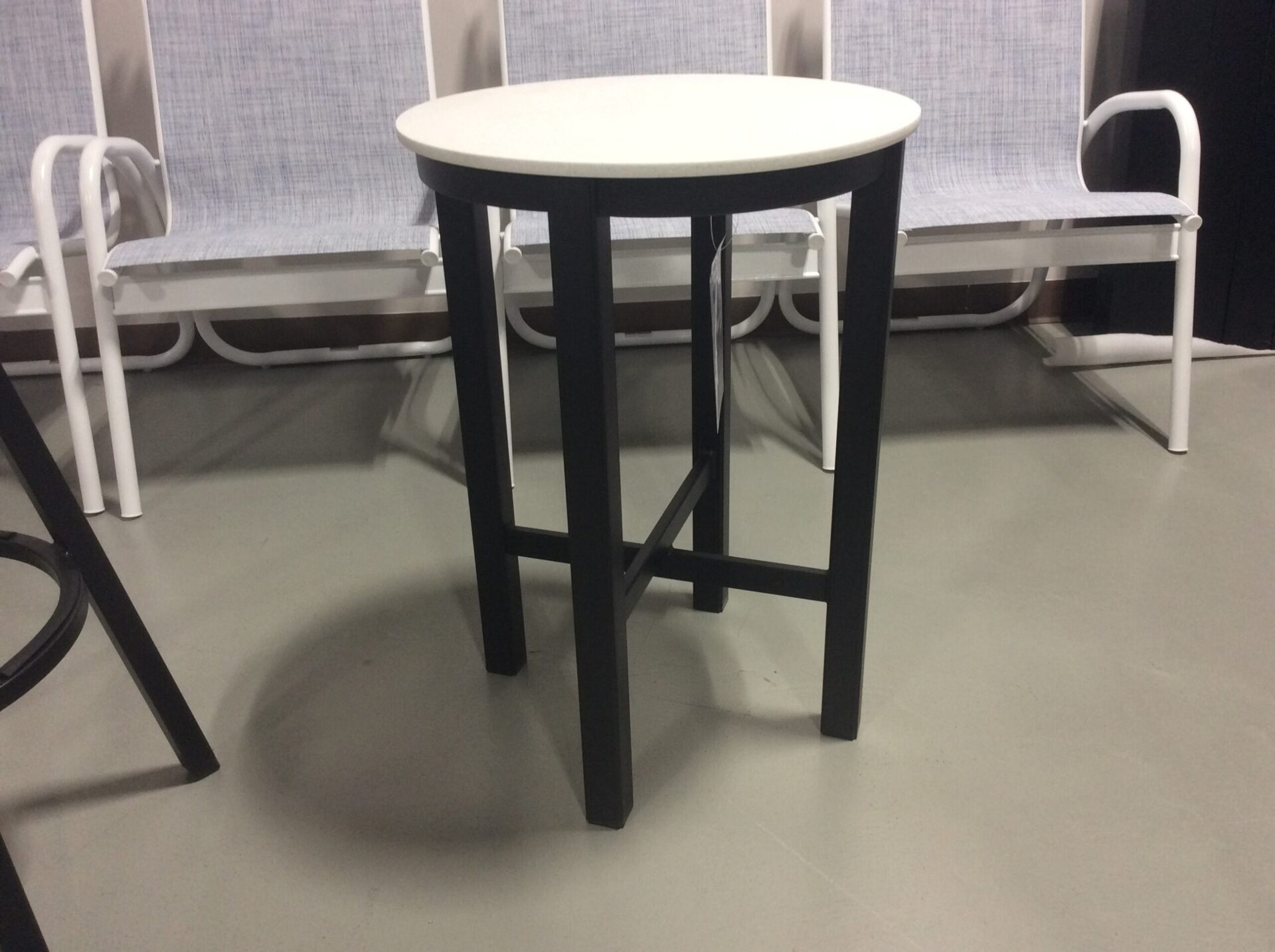 A small, round, white tabletop with black legs, standing in a room with white curtains and grey flooring. two white chairs are partially visible behind the table.