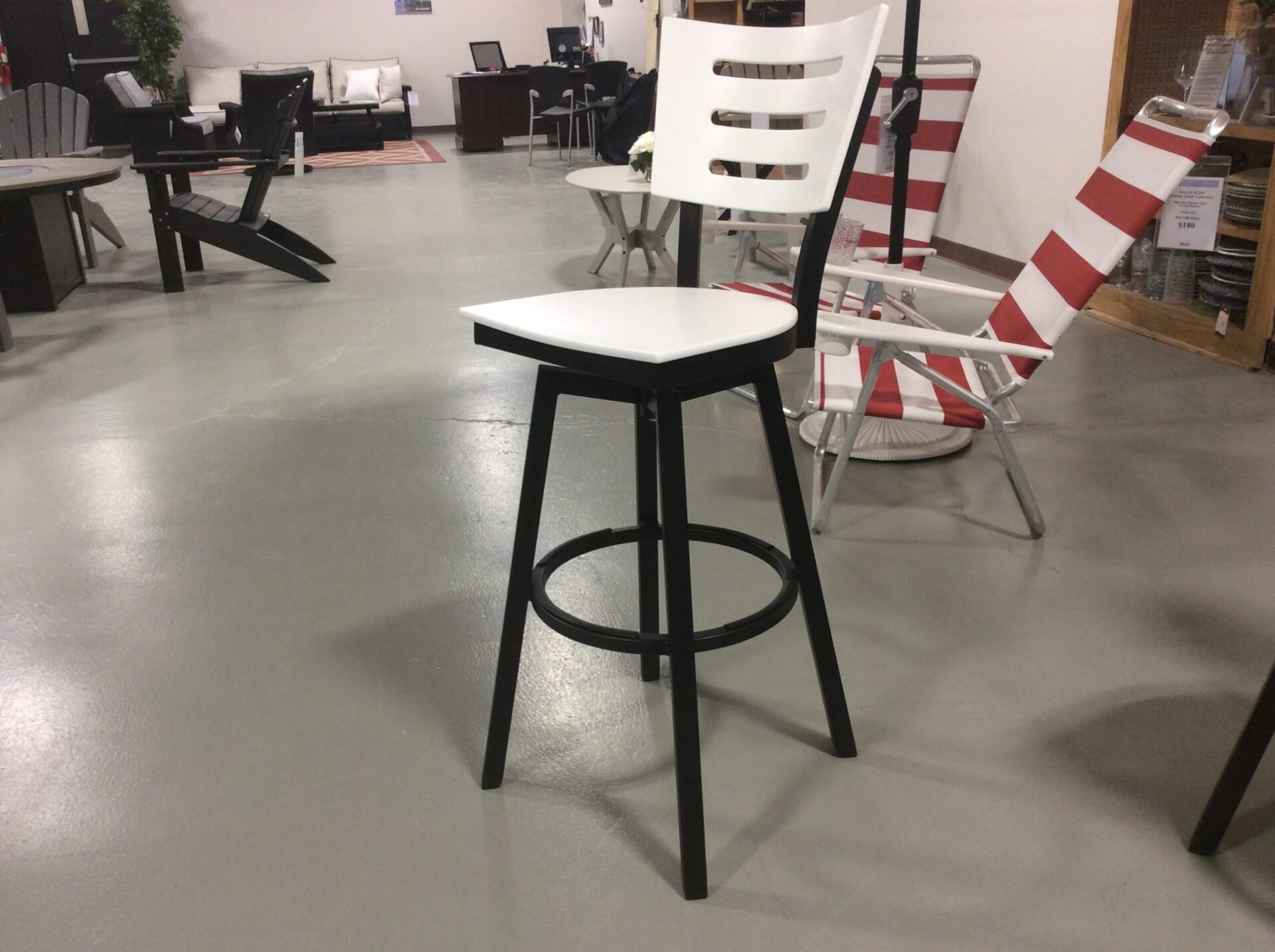 A white bar stool with a black frame is displayed in a spacious room featuring various furniture items, including chairs and tables on a polished concrete floor.