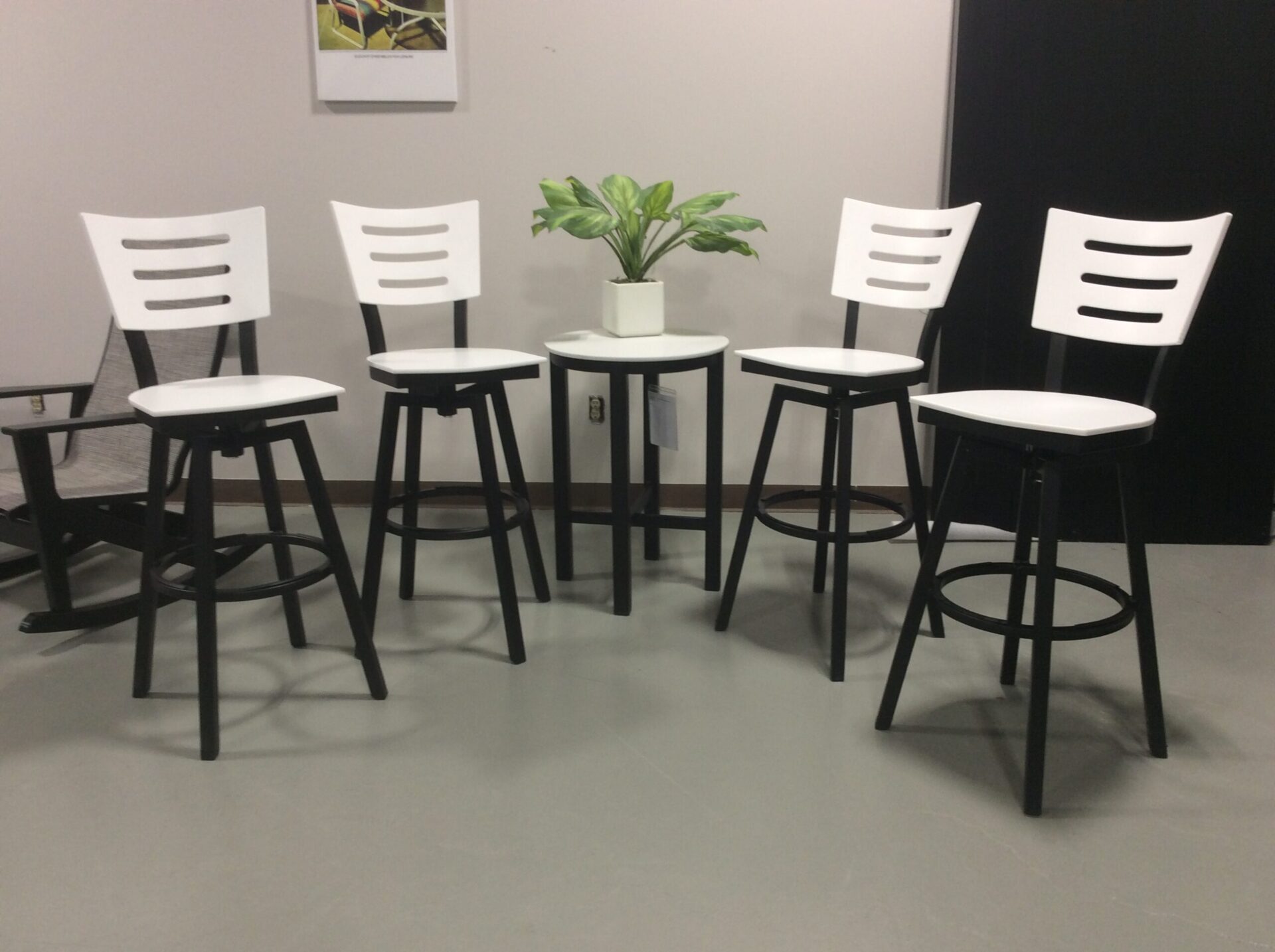 Four white modern bar stools arranged around a small white table with a green potted plant in the center, on a gray floor in a simple interior setting.