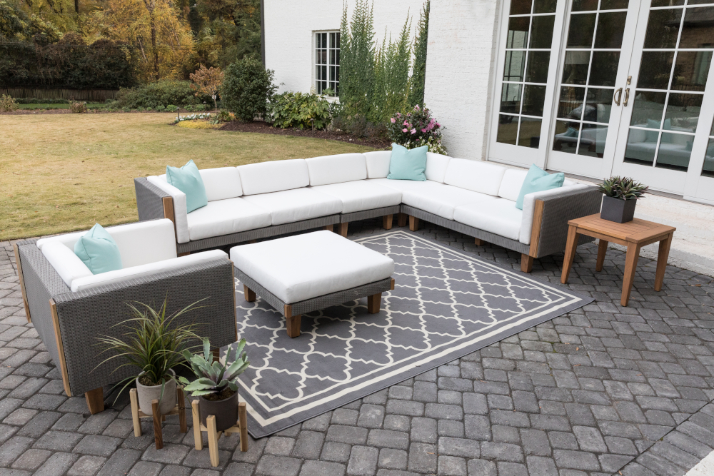 Elegant outdoor patio furniture arrangement featuring a light gray Catalina sectional sofa, matching chairs and ottoman, set on a textured rug, with a wooden coffee table, all surrounded by a lush garden.