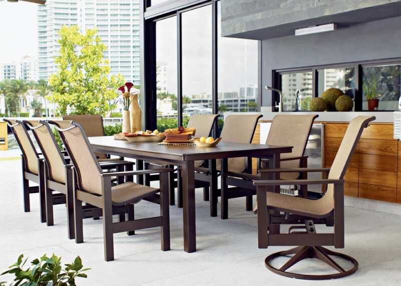A modern outdoor dining area on a balcony with a wooden table set for eight, sleek chairs, and urban skyscraper views in the background.