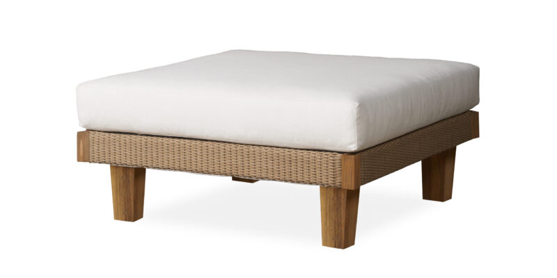 A minimalist outdoor ottoman with a woven tan base and sturdy wooden legs, topped with a thick white cushion, isolated on a white background.