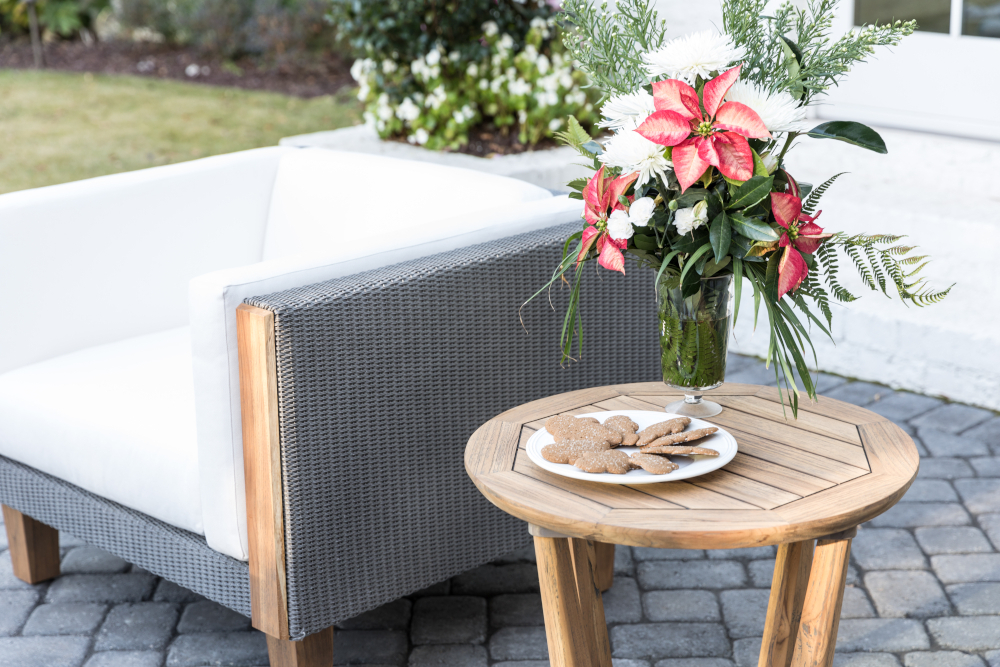 A modern outdoor patio setting featuring a stylish Catalina gray sofa with white cushions, a round wooden table with a plate of cookies, and a vase with red and white flowers.