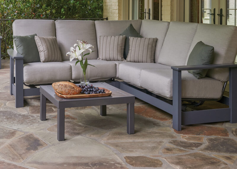 An outdoor seating area featuring a gray sectional sofa with cushions and a small rectangular table with a tray of bread and grapes, and a vase with white lilies.