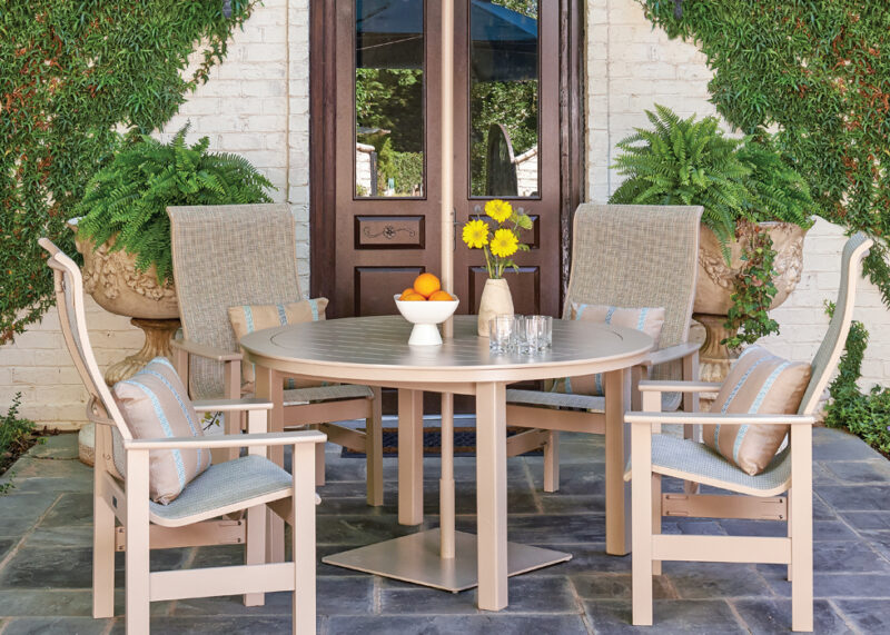An outdoor dining set with a round table and four chairs on a patio, surrounded by green climbing plants and a brick wall. the table is set with a bowl of oranges and a vase of yellow flowers.