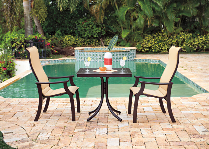 Two patio chairs and a table set with drinks by a pool, surrounded by lush greenery and tall palm trees, evoking a serene tropical outdoor dining setting.