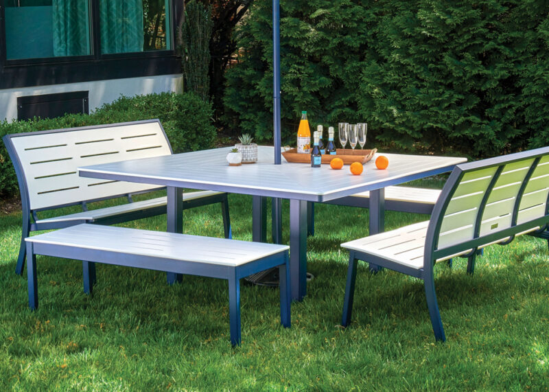 A modern outdoor dining set with a metallic blue table and matching benches, set on a lawn beside a house. the table is set with glasses, a bottle, and oranges.