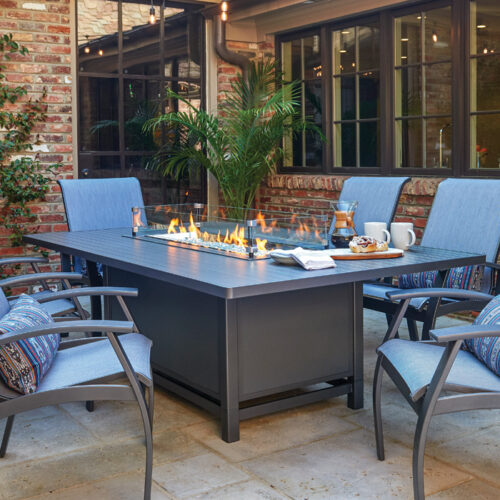 A cozy outdoor dining area with an aluminum slat top fire table, surrounded by cushioned chairs, set against a brick wall and green plants.