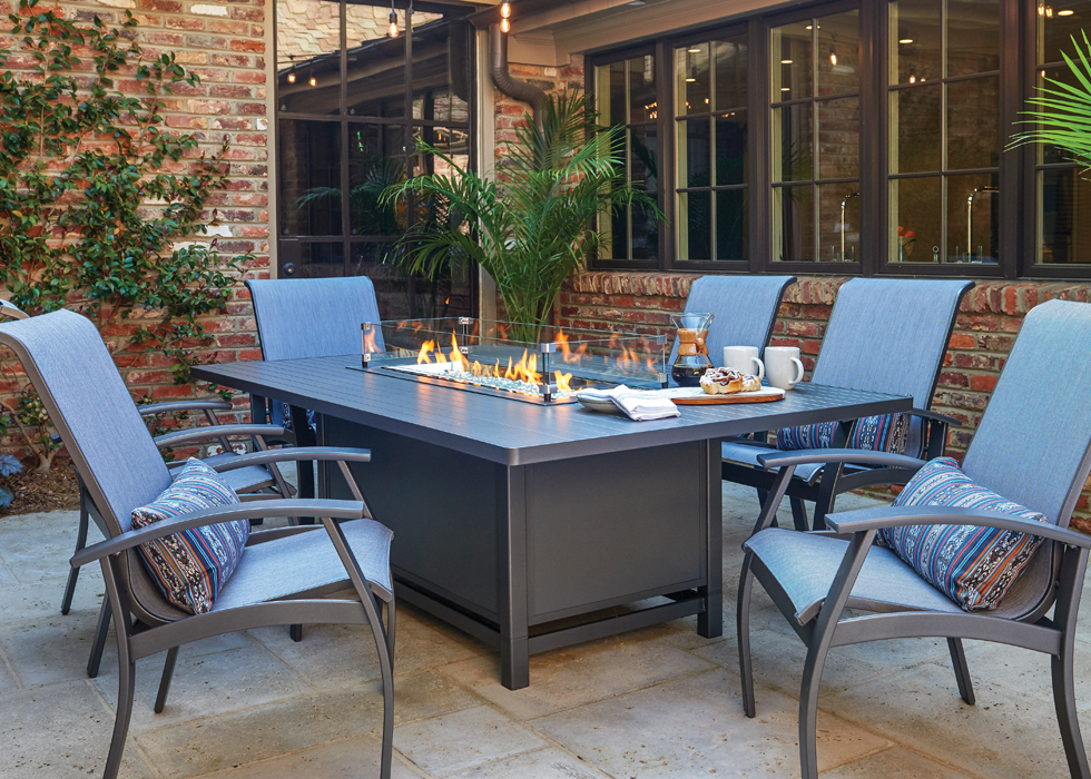 A cozy outdoor dining area with an aluminum slat top fire table, surrounded by cushioned chairs, set against a brick wall and green plants.