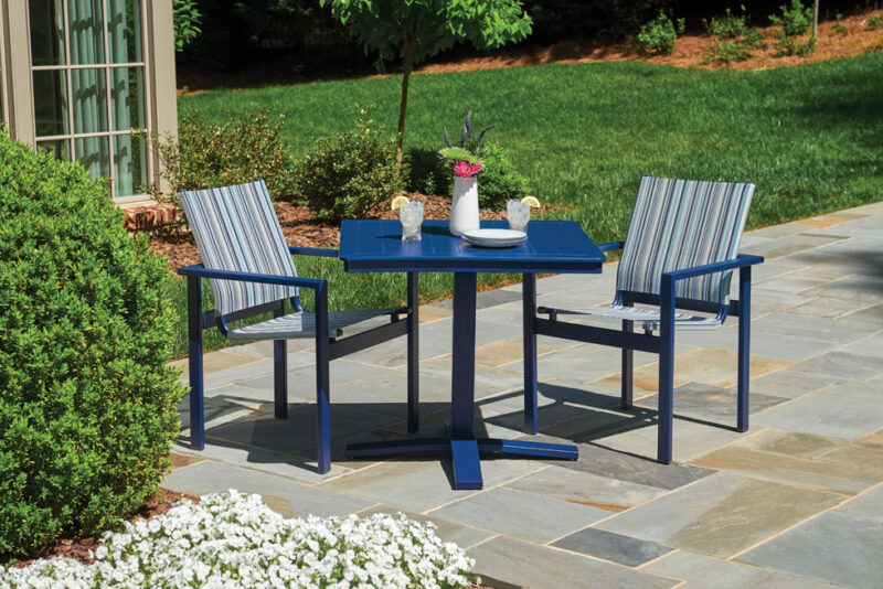 A serene outdoor setting featuring two blue-striped chairs and a matching blue table set with dishes and a vase of flowers, all positioned on a stone patio surrounded by lush greenery.