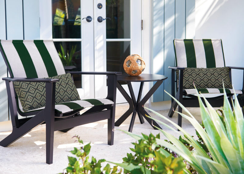 Two black patio chairs with green and white striped cushions beside a small round table, with a copper-coloured decorative sphere on it, set against a bright house entrance.