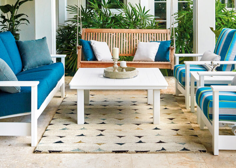 A stylish outdoor patio with a geometric rug, featuring a wooden bench, two armchairs with blue and white striped cushions, and a white coffee table, set against a lush backdrop.