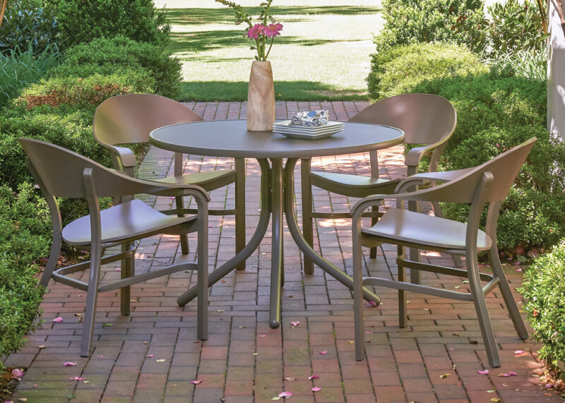 A serene outdoor setting with a round table and four chairs on a brick patio, surrounded by lush greenery. a vase with pink flowers and a book are on the table.