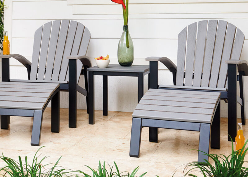 Two gray Adirondack chairs and matching footstools on a patio with a rustic polymer top table between them, holding a vase and a bowl of fruit.